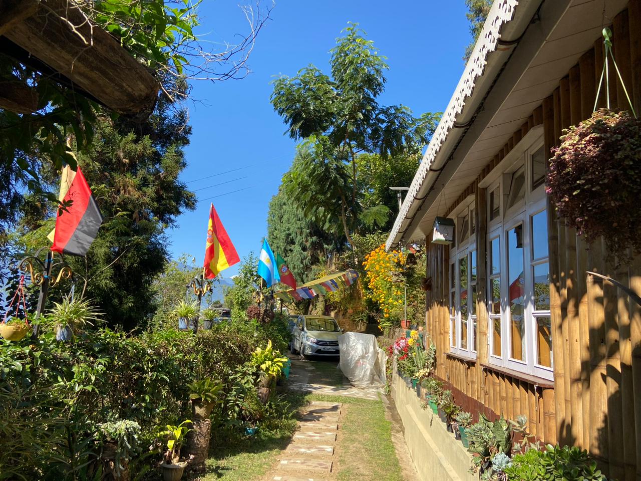 Exterior of Best HomestaysDarjeeling : cozy cottage nestled amidst greenery, surrounded by mountains against a clear sky backdrop.